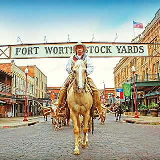 A cowboy at the entrance of the Fort Worth Stock Yards Photo: Visit Fort Worth A cowboy in customary western attire rides a brown and white horse underneath a "Fort Worth Stock Yards" sign that spans across a street lined with old western buildings